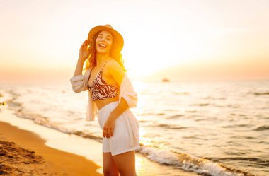 Sahilde günbatımının tadını çıkaran şapkalı güzel bir kadının portresi. Genç kadın özgürlüğü hissediyor, doğada rahatlıyor. Keyif ve rahatlama kavramı.