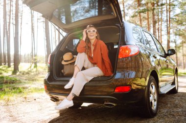 Neşeli kadın gün batımında ormanda bir arabanın bagajında oturur. Gezgin doğanın tadını çıkarır, özgürlüğü hisseder. Yolculuk, hafta sonu konsepti. Etkin yaşam biçimi.