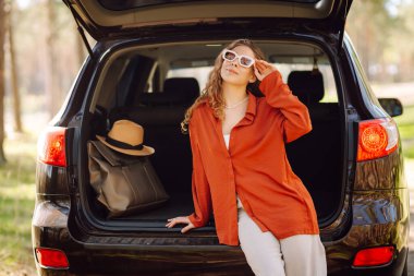 Neşeli kadın gün batımında ormanda bir arabanın bagajında oturur. Gezgin doğanın tadını çıkarır, özgürlüğü hisseder. Yolculuk, hafta sonu konsepti. Etkin yaşam biçimi.