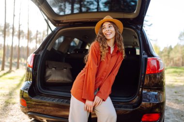 Neşeli kadın gün batımında ormanda bir arabanın bagajında oturur. Gezgin doğanın tadını çıkarır, özgürlüğü hisseder. Yolculuk, hafta sonu konsepti. Etkin yaşam biçimi.