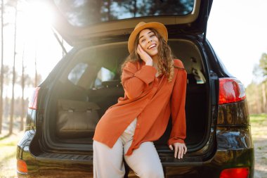 Neşeli kadın gün batımında ormanda bir arabanın bagajında oturur. Gezgin doğanın tadını çıkarır, özgürlüğü hisseder. Yolculuk, hafta sonu konsepti. Etkin yaşam biçimi.