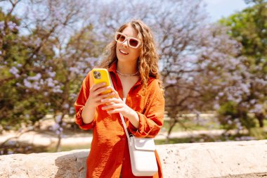 Parlak elbiseli, güneşli parkta telefonla yürüyen güzel bir kadın. Telefon blogları olan bir turist, tatilin tadını çıkarıyor. Yürüme ve eğlenme kavramı.