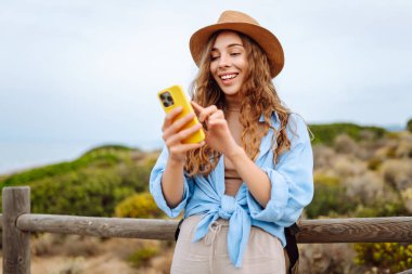 Yürüyen ahşap bir yolda telefonu ve şapkası olan sevimli bir turist blog yazıyor. Deniz manzarası seyreden güzel bir kadın. Etkin yaşam biçimi.