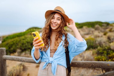 Yürüyen ahşap bir yolda telefonu ve şapkası olan sevimli bir turist blog yazıyor. Deniz manzarası seyreden güzel bir kadın. Etkin yaşam biçimi.