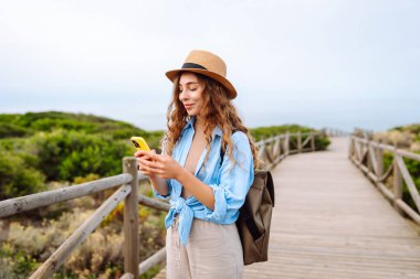 Yürüyen ahşap bir yolda telefonu ve şapkası olan sevimli bir turist blog yazıyor. Deniz manzarası seyreden güzel bir kadın. Etkin yaşam biçimi.