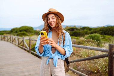 Yürüyen ahşap bir yolda telefonu ve şapkası olan sevimli bir turist blog yazıyor. Deniz manzarası seyreden güzel bir kadın. Etkin yaşam biçimi.