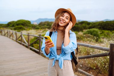 Yürüyen ahşap bir yolda telefonu ve şapkası olan sevimli bir turist blog yazıyor. Deniz manzarası seyreden güzel bir kadın. Etkin yaşam biçimi.