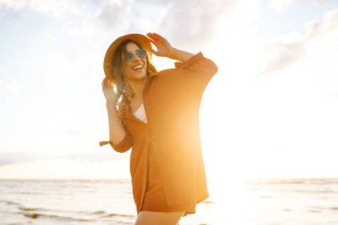 Şapkalı tatlı bir kadın özgürlüğünü hissediyor ve kumsalda günbatımının tadını çıkarıyor. Dışarıdan temiz hava soluyan şık elbiseli kadın portresi. Özgürlük, rahatlama ve zevk kavramı.