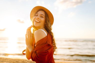 Şapkalı tatlı bir kadın özgürlüğünü hissediyor ve kumsalda günbatımının tadını çıkarıyor. Dışarıdan temiz hava soluyan şık elbiseli kadın portresi. Özgürlük, rahatlama ve zevk kavramı.