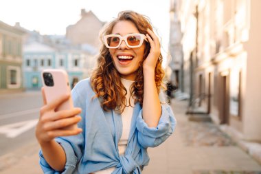 Gözlüklü ve telefonlu şık bir kadın şehirde eğleniyor. Genç bir kadın telefonda havanın ve mesajların tadını çıkarıyor. Turizm, güzellik konsepti.