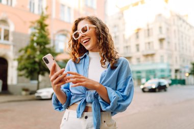 Gözlüklü ve telefonlu şık bir kadın şehirde eğleniyor. Genç bir kadın telefonda havanın ve mesajların tadını çıkarıyor. Turizm, güzellik konsepti.