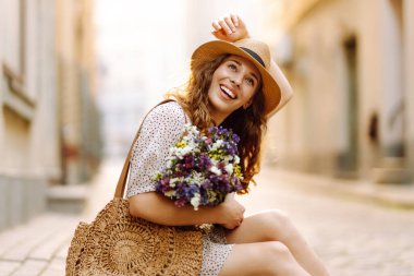 Elinde güzel bir buket çiçekle sevimli bir kadın Avrupa caddesinde günbatımının tadını çıkarıyor. Şapkalı güzel bir kadın dışarıda dinleniyor. Tatil ve güzellik konsepti.