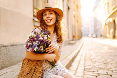 Elinde güzel bir buket çiçekle sevimli bir kadın Avrupa caddesinde günbatımının tadını çıkarıyor. Şapkalı güzel bir kadın dışarıda dinleniyor. Tatil ve güzellik konsepti.