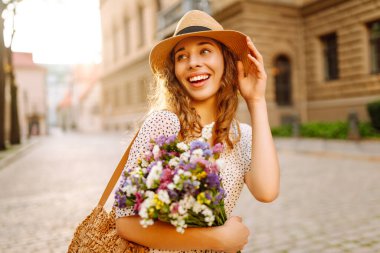 Elinde güzel bir buket çiçekle sevimli bir kadın Avrupa caddesinde günbatımının tadını çıkarıyor. Şapkalı güzel bir kadın dışarıda dinleniyor. Tatil ve güzellik konsepti.