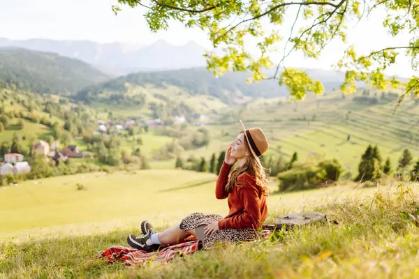 Çimenli yamaçta oturan genç bir kadının arka görüntüsü. Şapkalı gezgin dağ manzarasının ve doğada gün batımının tadını çıkarıyor. Özgürlük kavramı. Etkin yaşam biçimi.