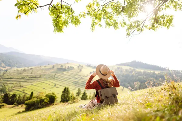 Çimenli yamaçta oturan genç bir kadının arka görüntüsü. Şapkalı gezgin dağ manzarasının ve doğada gün batımının tadını çıkarıyor. Özgürlük kavramı. Etkin yaşam biçimi.