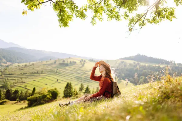 Çimenli yamaçta oturan genç bir kadının arka görüntüsü. Şapkalı gezgin dağ manzarasının ve doğada gün batımının tadını çıkarıyor. Özgürlük kavramı. Etkin yaşam biçimi.