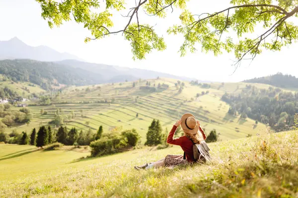 Çimenli yamaçta oturan genç bir kadının arka görüntüsü. Şapkalı gezgin dağ manzarasının ve doğada gün batımının tadını çıkarıyor. Özgürlük kavramı. Etkin yaşam biçimi.