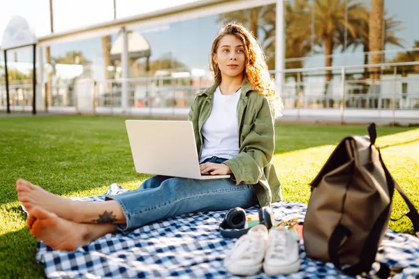 Genç bir kadın gün batımında yeşil bir çayırda dizüstü bilgisayarla çalışıyor. Kadın serbest çalışan, parktaki bir paspasın üzerinde palmiye ağaçlarının arka planında oturuyor. Serbest çalışma, blog yazma, rahatlama kavramı.