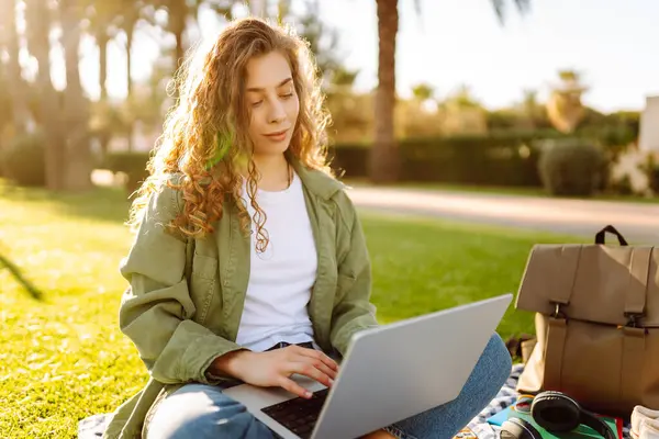 Genç bir kadın gün batımında yeşil bir çayırda dizüstü bilgisayarla çalışıyor. Kadın serbest çalışan, parktaki bir paspasın üzerinde palmiye ağaçlarının arka planında oturuyor. Serbest çalışma, blog yazma, rahatlama kavramı.