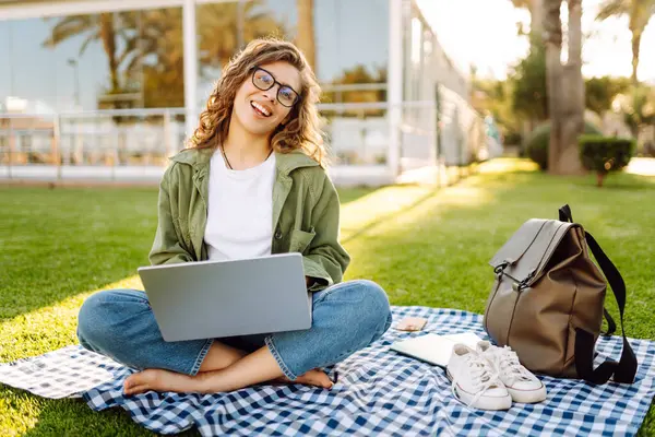 Genç bir kadın gün batımında yeşil bir çayırda dizüstü bilgisayarla çalışıyor. Kadın serbest çalışan, parktaki bir paspasın üzerinde palmiye ağaçlarının arka planında oturuyor. Serbest çalışma, blog yazma, rahatlama kavramı.