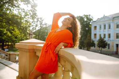 Parlak elbiseli güzel bir kadın gün batımında güzel bir binanın yanında poz veriyor. Mutlu kadın güneşin tadını çıkarıyor. Güzellik, moda ve eğlence konsepti.
