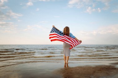 Neşeli bir kadın gün batımında Amerikan bayrağıyla kumsalda yürürken eğlenir. Genç bir kadın mutlu ve Amerikan bayrağıyla doğanın tadını çıkarıyor. Özgürlük ve keyif kavramı.