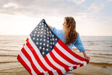Neşeli bir kadın gün batımında Amerikan bayrağıyla kumsalda yürürken eğlenir. Genç bir kadın mutlu ve Amerikan bayrağıyla doğanın tadını çıkarıyor. Özgürlük ve keyif kavramı.