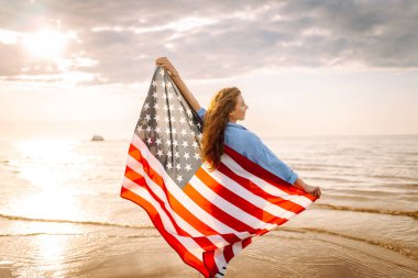 Neşeli bir kadın gün batımında Amerikan bayrağıyla kumsalda yürürken eğlenir. Genç bir kadın mutlu ve Amerikan bayrağıyla doğanın tadını çıkarıyor. Özgürlük ve keyif kavramı.