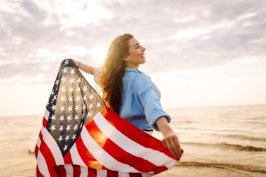 Neşeli bir kadın gün batımında Amerikan bayrağıyla kumsalda yürürken eğlenir. Genç bir kadın mutlu ve Amerikan bayrağıyla doğanın tadını çıkarıyor. Özgürlük ve keyif kavramı.