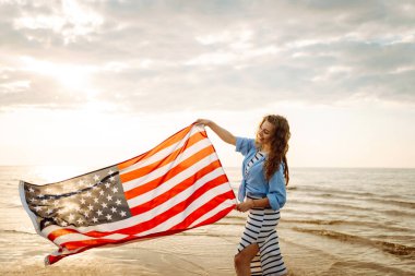 Neşeli bir kadın gün batımında Amerikan bayrağıyla kumsalda yürürken eğlenir. Genç bir kadın mutlu ve Amerikan bayrağıyla doğanın tadını çıkarıyor. Özgürlük ve keyif kavramı.