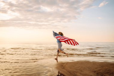 Neşeli bir kadın gün batımında Amerikan bayrağıyla kumsalda yürürken eğlenir. Genç bir kadın mutlu ve Amerikan bayrağıyla doğanın tadını çıkarıyor. Özgürlük ve keyif kavramı.