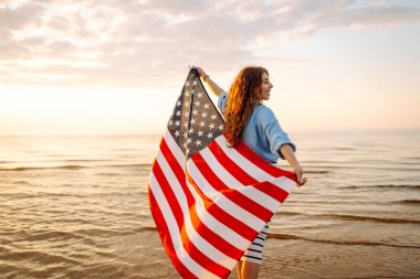 Neşeli bir kadın gün batımında Amerikan bayrağıyla kumsalda yürürken eğlenir. Genç bir kadın mutlu ve Amerikan bayrağıyla doğanın tadını çıkarıyor. Özgürlük ve keyif kavramı.