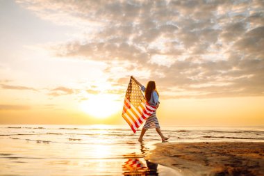 Neşeli bir kadın gün batımında Amerikan bayrağıyla kumsalda yürürken eğlenir. Genç bir kadın mutlu ve Amerikan bayrağıyla doğanın tadını çıkarıyor. Özgürlük ve keyif kavramı.