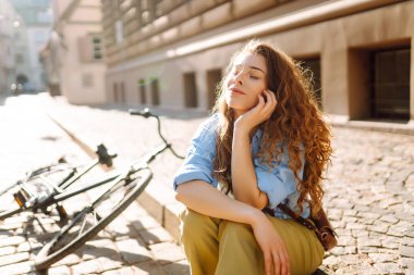 Güneşli bir caddede oturan kıvırcık saçlı genç bir kadının portresi. Gün batımında bisiklet sürdükten sonra dinlenen güzel bir turist. Binicilik kavramı, dinlenme. Etkin yaşam biçimi.