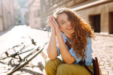 Güneşli bir caddede oturan kıvırcık saçlı genç bir kadının portresi. Gün batımında bisiklet sürdükten sonra dinlenen güzel bir turist. Binicilik kavramı, dinlenme. Etkin yaşam biçimi.