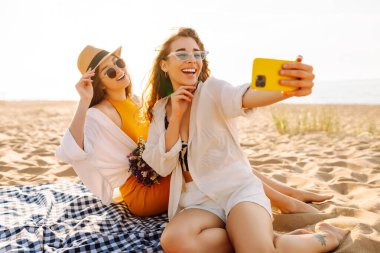 Bir buket çiçekli iki mutlu kadın kumlu bir sahilde selfie için poz veriyor. Güzel arkadaşlar gün batımında pikniğin ve havanın tadını çıkarıp telefonlarıyla fotoğraf çekerler. Blog yazma, boş zaman kavramı.