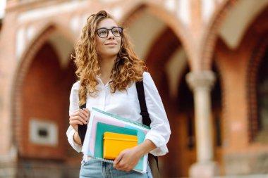 Kıvırcık saçlı ve gözlüklü genç bir öğrenci tarihi üniversite geçmişine karşı elinde kitap ve defterler tutuyor. Güzel bir kadın üniversite kampüsünün yakınında dinleniyor. Eğitim kavramı.