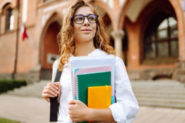 Kıvırcık saçlı ve gözlüklü genç bir öğrenci tarihi üniversite geçmişine karşı elinde kitap ve defterler tutuyor. Güzel bir kadın üniversite kampüsünün yakınında dinleniyor. Eğitim kavramı.
