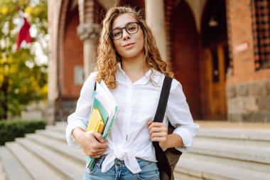 Kıvırcık saçlı ve gözlüklü genç bir öğrenci tarihi üniversite geçmişine karşı elinde kitap ve defterler tutuyor. Güzel bir kadın üniversite kampüsünün yakınında dinleniyor. Eğitim kavramı.