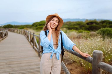 Şapkalı güzel bir bayan turist deniz manzaralı ahşap bir yolda yürüyor. Macerayı seven ve gün doğumunda yürüyen genç bir kadın. Seyahat, tatil konsepti. Etkin yaşam biçimi.