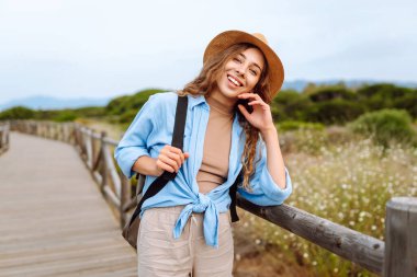 Şapkalı güzel bir bayan turist deniz manzaralı ahşap bir yolda yürüyor. Macerayı seven ve gün doğumunda yürüyen genç bir kadın. Seyahat, tatil konsepti. Etkin yaşam biçimi.