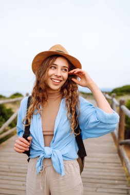Şapkalı güzel bir bayan turist deniz manzaralı ahşap bir yolda yürüyor. Macerayı seven ve gün doğumunda yürüyen genç bir kadın. Seyahat, tatil konsepti. Etkin yaşam biçimi.