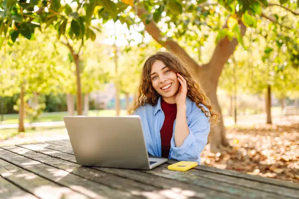 Güneşli bir parktaki ahşap bir masada çalışan dizüstü bilgisayarı olan şık bir kadın. Açık havada çalışan güzel bir bayan serbest yazar. Uzaktan çalışma, eğlence ve eğitim kavramı.