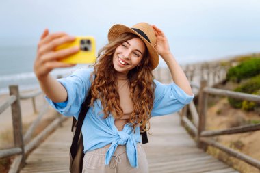 Yürüyüş parkurunda sırt çantalı neşeli bir kadın telefonda selfie çekiyor. Güzel bayan gezgin gündoğumunda doğanın tadını çıkarıyor. Macera, blog konsepti.