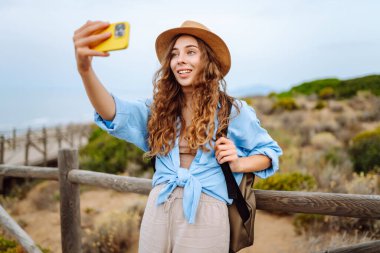 Yürüyüş parkurunda sırt çantalı neşeli bir kadın telefonda selfie çekiyor. Güzel bayan gezgin gündoğumunda doğanın tadını çıkarıyor. Macera, blog konsepti.