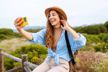 Yürüyüş parkurunda sırt çantalı neşeli bir kadın telefonda selfie çekiyor. Güzel bayan gezgin gündoğumunda doğanın tadını çıkarıyor. Macera, blog konsepti.