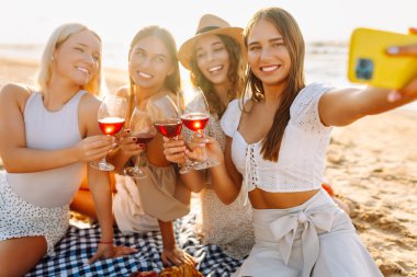 Neşeli kız arkadaşlar sahilde rahatlar, telefonda selfie çeker ve bardaktan içki içerler. Dört kadın deniz kıyısında pikniğin tadını çıkarıyor. Dostluk kavramı, kutlama.