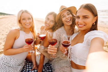 Neşeli kız arkadaşlar sahilde rahatlar, telefonda selfie çeker ve bardaktan içki içerler. Dört kadın deniz kıyısında pikniğin tadını çıkarıyor. Dostluk kavramı, kutlama.