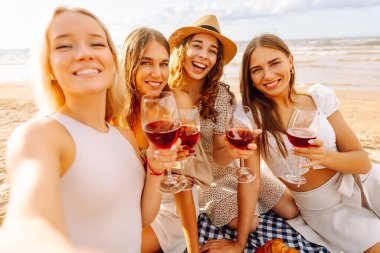 Neşeli kız arkadaşlar sahilde rahatlar, telefonda selfie çeker ve bardaktan içki içerler. Dört kadın deniz kıyısında pikniğin tadını çıkarıyor. Dostluk kavramı, kutlama.